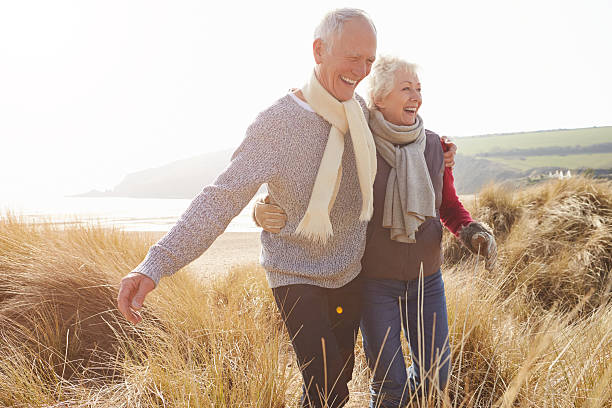 coppia senior a piedi attraverso dune di sabbia sulla spiaggia d'inverno - holding hands couple senior couple togetherness foto e immagini stock