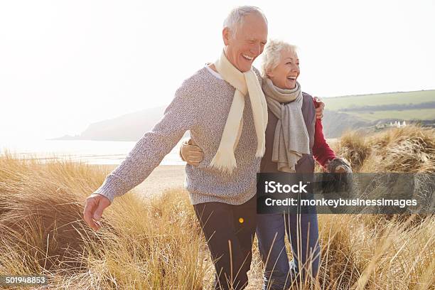 Senior Couple Walking Through Sand Dunes On Winter Beach Stock Photo - Download Image Now