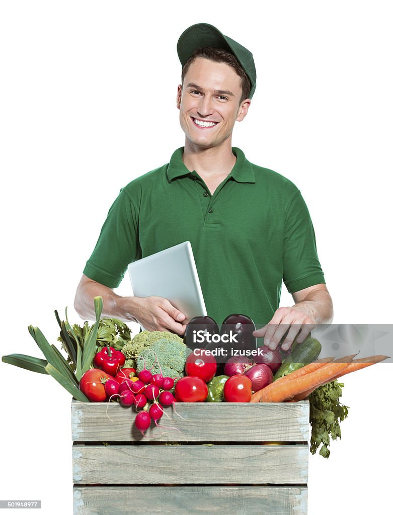 Delivery man with organic food Delivery man delivering box with organic food, holding a digital tablet and smiling  at camera. Studio shot, white background. Adult Stock Photo