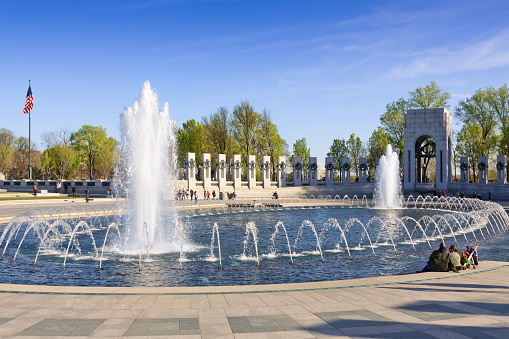 Washington DC, USA - March 27, 2012: Fountains at the World War II Memorial in Washington DC. Young interracial family with a stroller is resting by the fountain. Green trees, pool, American flag and deep blue clear sky are in the image. Canon EF 24-105mm/4L IS USM Lens.