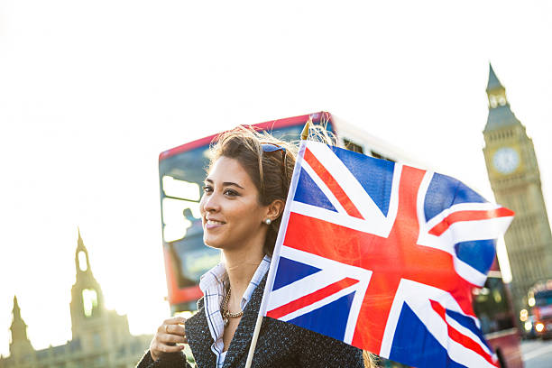 London - happy tourist holding British UK flag by Big Ben and red double decker bus. Excited girl sightseeing travel on Westminster Bridge, London, England, United Kingdom. Multiracial Asian Caucasian