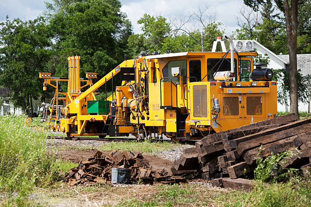 A railroad construction site with old wood ties piled in the right foreground. The background machine is a ballast tamper which aligns the tracks and packs ballast (rock) under the tracks to make them level and parallel for durability and a smooth ride.