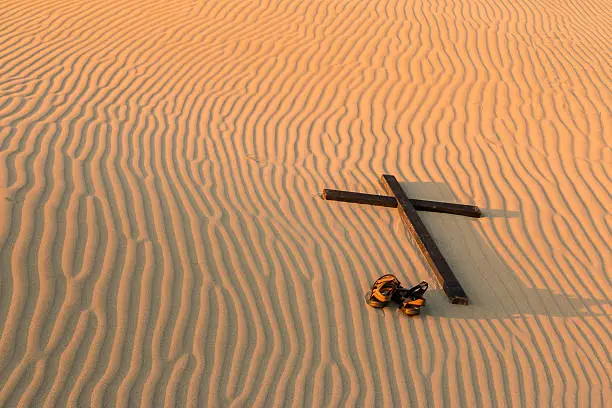 Pair of sandles by a black cross on waves of sand.