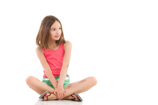Young girl sitting on the floor with legs crossed and looking away. Full length studio shot isolated on white.