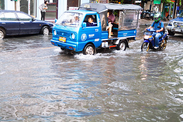 Bangkok, Thailand - May 6, 2014: Street and traffic scene in Bangkok with high water after short and heavy tropical rain shower. A local tuk-tuk is driving through water, a motorcycle is following. Inside of tuk-tuk are passengers, one is sitting next to driver in front. Sunset shot. A woman is standing in background n sidewalk.