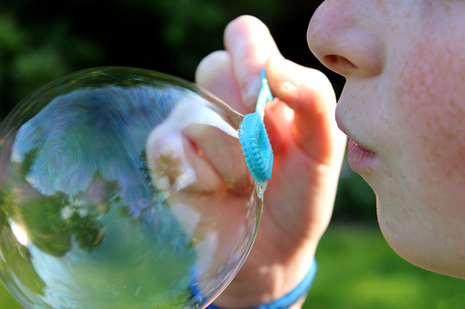 Photo showing a young girl blowing bubbles in the garden.  She is pictured blowing a particularly large bubble, which is lit up by the afternoon sunshine, causing reflections and rainbow colours to appear on the surface of the bubble, rather like oil.