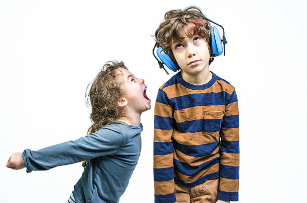 Young girl yelling at brother wearing ear protection Young girl yelling at brother wearing ear protection while brother is kind of / bored of it and is looking up and sulking, studio shot rolling eyes irritation displeased facial expression stock pictures, royalty-free photos & images