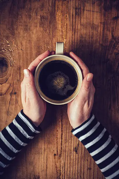 Lonely woman drinking coffee in the morning, top view of female hands holding cup of hot beverage on wooden desk, retro toned.