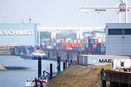 Duisburg, Germany - July 12, 2014: Tele distance shot of inland port Duisburg in Ruhrgebiet. Freight ship is anchored at pier. On pier are many cargo containers. Above containers is hge crane. At left side is huge hangar. View over water and along piers.