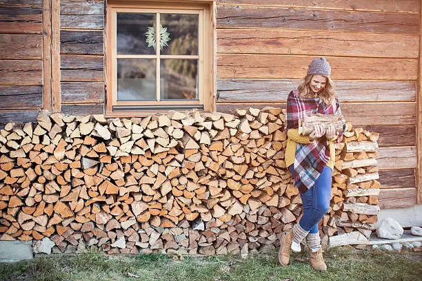 Woman on vacation at mountain cabin. Standing in front of house and enjoying in winter day. Carrying stack of wood for fire.  Wearing warm clothing, hat and scarf. Austrian Alps.
