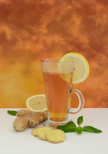 Glass of freshly made hot ginger tea with a slice of lemon. Surrounding it are other raw ingredients of mint leaves, fresh lemon and chopped slices of ginger.