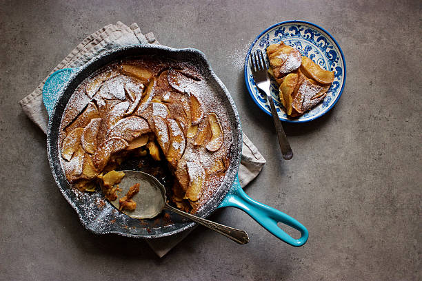 Roasted apple clafoutis (French custard cake) in cast iron pan A homemade French dessert: a clafoutis, a French-style custard cake, filled with roasted apples, that has been baked in the oven then placed on top of a tea towel on a grey kitchen countertop and sprinkled with icing sugar. One slice has been removed to a blue and white plate with a fork and a vintage silver serving tool is resting in the pan. clafoutis stock pictures, royalty-free photos & images