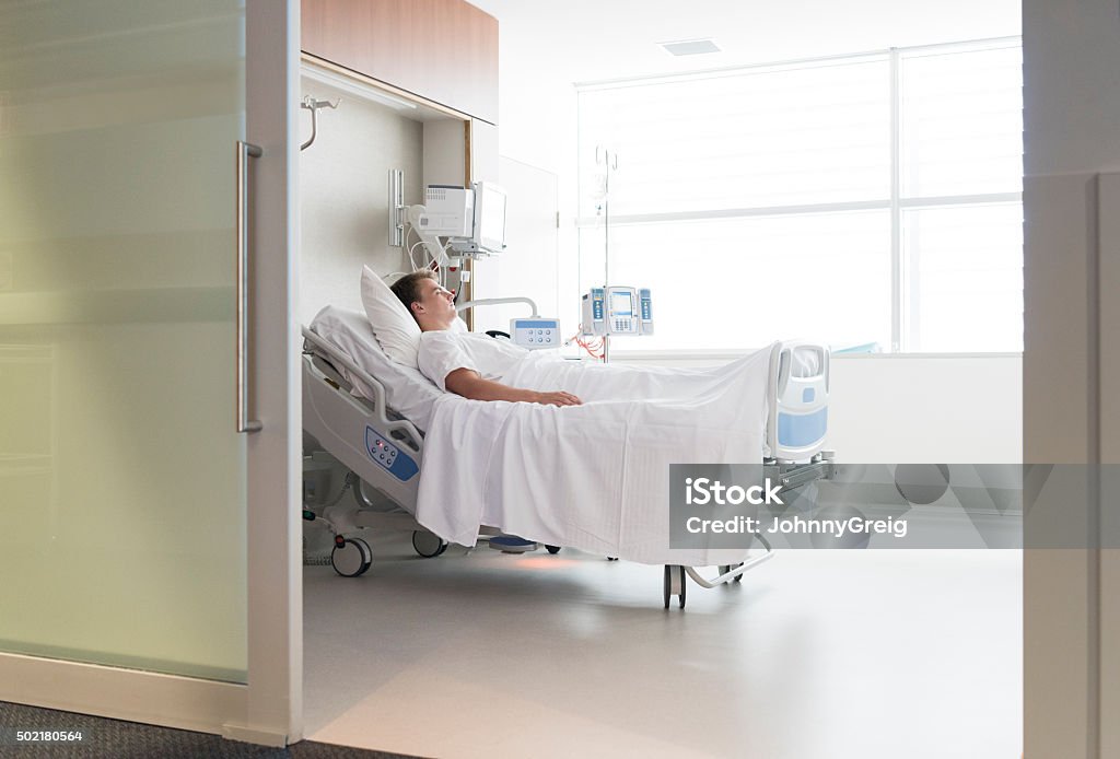 Male patient lying in hospital bed, view through doorway Young man resting on hospital ward. Illness, health, recovery, rehabilitation. He is lying down in a bed in private room  looking thoughtfully out of the window. Patient Stock Photo