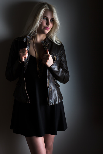 Beautiful teenage fashion model leaning against wall and posing in black dress with leather jacket.