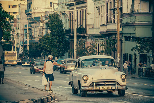 Havana, Cuba-November 7, 2015. Busy Boulevard Calzada De Infanta in Centro Havana. Old, rundown residential and commercial buildings, remembering better times from 50 years ago. Street life in full effect: lot of people on the street. Pedestrians in the background. Man stopping the taxi on the curb (private old American cars, local version of \