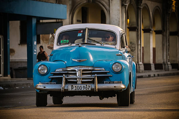Havana, Cuba - November 7, 2015. Old American made car  driving along Havana Boulevard, Cuba.  Diver looking ahead. Passengers sharing the space. City architecture in the background.  One of countless vintage US made cars imported to Cuba before the Revolution but still running after more than 50 years due to ingenuity of Cuban car mechanics and some help of various old car parts, including the left over of Russian diesel engines. Most of these cars run across the city as TAXI for local Cubans. Some cars, that are restored completely and looking as new are in use now for foreign tourists only, with hourly rate of $25 per hour.