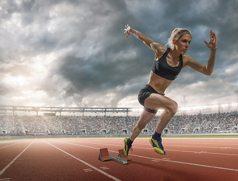 A low angle view of a woman sprinter in mid motion exploding from the starting blocks in an outdoor running track in preparation for a race. The young athlete is  wearing generic black running knickers and top and is competing in a floodlit outdoor athletics track under a stormy evening sky. 