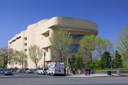 Washington DC, USA- March 26, 2012: The National Museum of the American Indian, Washington DC, USA. It is part of the Smithsonian Institution and is dedicated to the life, languages, literature, history, and arts of the Native Americans. Green trees, street, sidewalk, parked cars, traffic light, street food vendor truck, passersby and vivid blue clear sky are in the image. Canon EF 24-105mm/4L IS USM Lens.