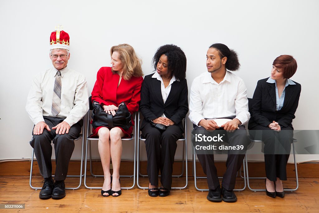 Best Man for the Job A group of business colleagues sitting in a waiting room for an interview. One is wearing a crown. Male/white privilege concept. Employment And Labor Stock Photo