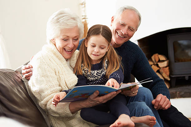 бабушкой и дедушкой и внучка читать книгу вместе дома - grandparent family reading inside of стоковые фото и изображения