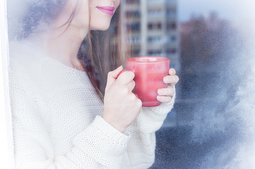 Beautiful young woman looking through the window on a winter day. She is holding a cup of tea, in warm clothes. Ice visible on the glass in the foreground. Indoor shot.