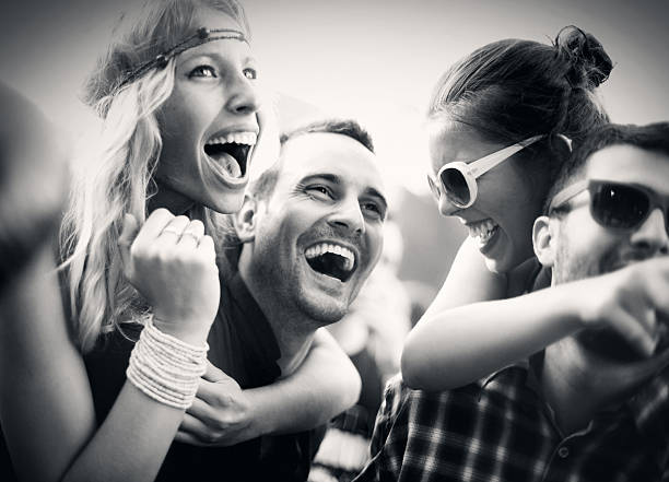 Group of young adults at a concert.Two men carrying their girls on shoulders.Girls are ecstatic and having big fun while looking at stage and laughing.It's summer sunset.Large crowd in background,blurry.Low angle shot. Black and white image.