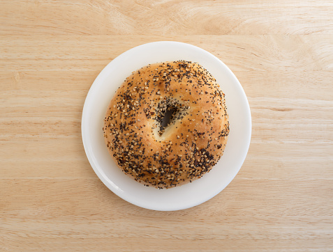 Top view of a bagel with several different types of seasonings on a white plate atop a wood table top.