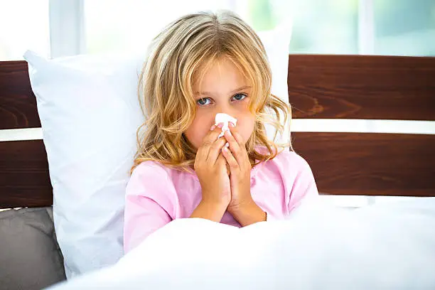 Photo of little girl sitting on white bed in morning. Girl having runny nose