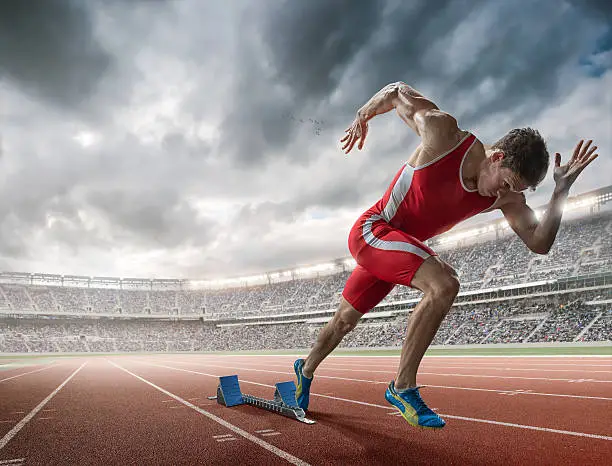 A mid action image of a professional male athlete sprinting from starting blocks on an outdoor athletics running track. The runner is in a generic outdoor floodlit stadium full of spectators under a dramatic stormy sky. The sprinter wears a red body suit and running spikes during a practice session. 