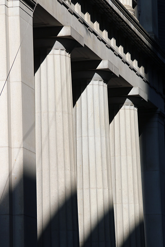 A vertical, close-up image of legal pillars.  There are four columns with vertical lines running through them and sharp, triangular black shadows at the top and bottom.  There are some horizontal lines running above the pillars, which also cast dark, contrasted shadows.