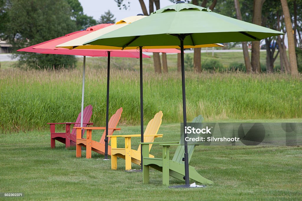 Colorful adirondack chairs in a row. A row of chairs with matching umbrellas are in a line on a very large lawn.  Adirondack Chair Stock Photo