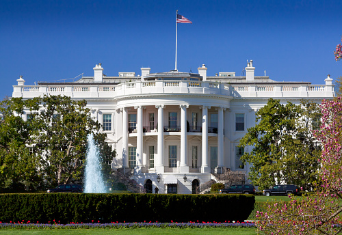 The South Portico of the White House. Washington DC. The White House is the official residence and principal workplace of the President of the United States, located at 1600 Pennsylvania Avenue. Beautifully landscaped lawn with flowers, fountain and blooming trees is in foreground. Deep blue clear sky is in background. American flag is flying atop. The image lit by spring evening sun. Canon 24-105mm f/4L lens.