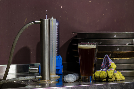 A messy brewers workbench with a hydrometer, thermometer, glass of beer and work gloves