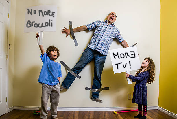 Kids rebellion led to strapping the father on wall A son and a daughter have hung their father on the wall with duct tape and keep him as a hostage while they are holding placards saying "More TV" and  "No more Broccoli" protest photos stock pictures, royalty-free photos & images