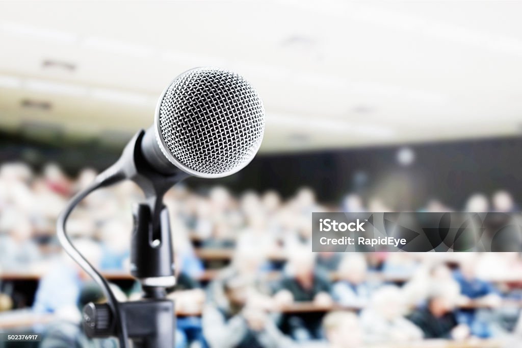 Large audience in auditorium with vocal mic await speaker A vocal microphone stands ready against a background of a large out-of-focus and unrecognizable audience at a performance, lecture, or meeting. Audience Stock Photo