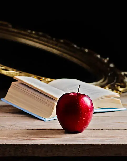 An red apple, a mirror and a book on wooden table. These elements tell the story of Snow White.