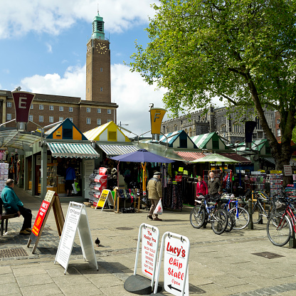 Norwich, Norfolk, England - May 13, 2014: People shopping along Gentleman’s Walk and in the Market Place in Norwich, England. Norwich Market has over 190 stalls and is famous worldwide for its brightly striped awnings (replaced in 2005 with fixed roofs). It is the largest Monday-Saturday market in the UK and there has been a market on the site since Norman times. Norwich City Hall and the Guildhall are in the background.