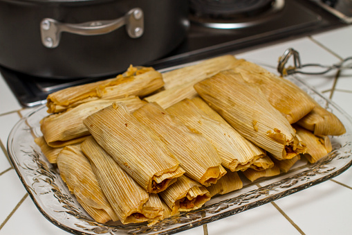 Pile of Ready to Eat Steamed Tamales on a Glass Serving Plate in a Kitchen next to a Cooking Pot with Tongs