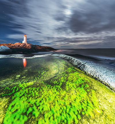 A puddle filled with a bright green algae is illuminated in the foreground of Peggy's Cove Lighthouse on the Atlantic coast of Nova Scotia.  Long exposure with light painting.
