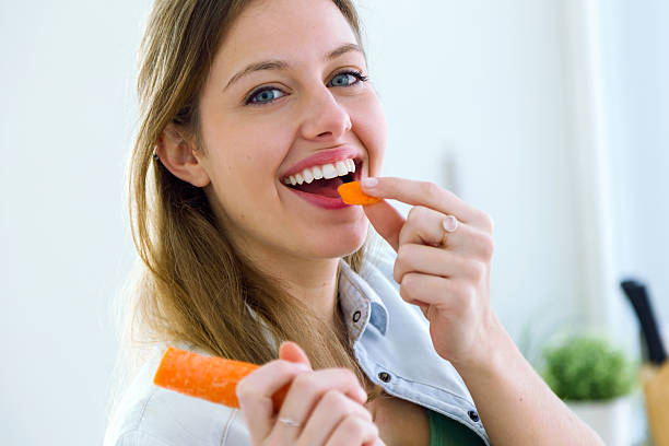 bastante joven mujer comiendo zanahoria en la cocina. - carrot fotografías e imágenes de stock