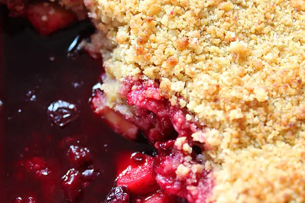 Photo showing a delicious summer fruit crumble, just taken out of the oven served up in a dish, as a generous portion.  The fruit in the crumble include chopped apple, raspberries, blackberries, blueberries, cherries, redcurrants and blackcurrants.