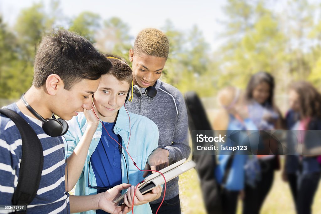 Music: Teenage friends listen to music on digital tablet.  Park. Multi-ethnic group of Jr. High school friends hang out on school campus or park. They listen to music with headphones on digital tablet. Girls in background.  Music Stock Photo