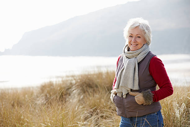 senior frau zu fuß auf sand dunes on winter beach - healthy lifestyle women beach looking at camera stock-fotos und bilder