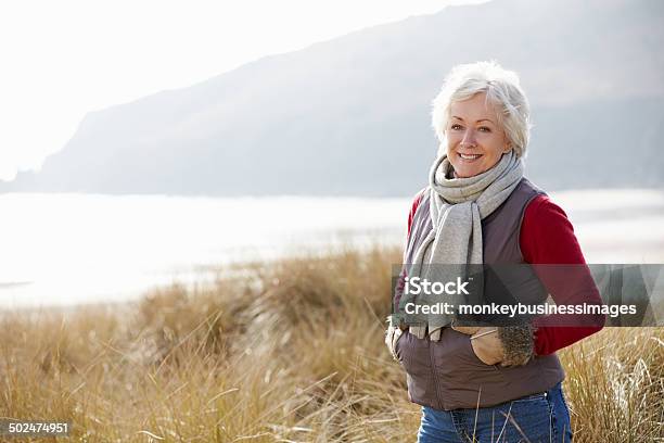 Senior Mujer Caminando A Través De Las Dunas De Arena En La Playa De Invierno Foto de stock y más banco de imágenes de Mujeres mayores