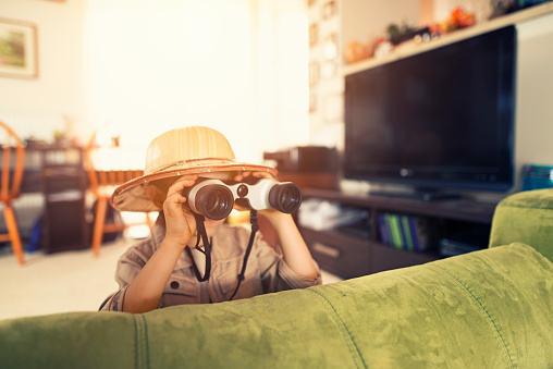 Little boy with binoculars exploring living room. The boy aged 6 is playing indoors safari exploration trip. In his imagination he is in the middle of dangerous jungle.