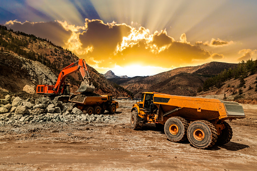 Excavator loading dumper trucks at sunset at mining site.