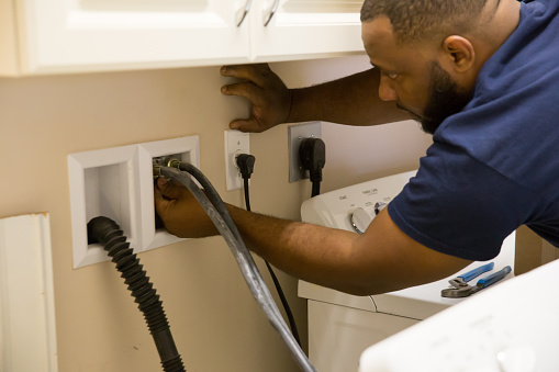 African American worker is in the laundry room of a home. He is removing the hoses attached to a washing machine that is being replaced with a new machine. He is unattaching the water lines from hook up on the wall. Shot taken with a Canon 5D Mark 3.  rm