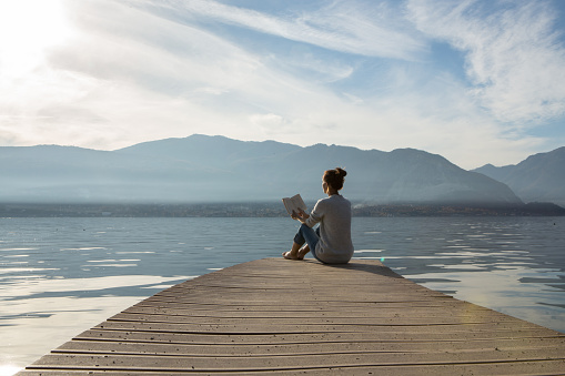 Young woman sits on a jetty above the lake, she is reading a book. Beautiful Autumn day in Italy.