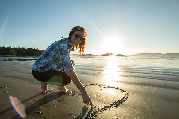 Young woman on beach drawing heart shape on sand-sunset