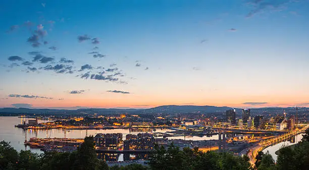 Blue chrome skies and orange glow of midsummer sunset over the iconic landmarks and waterfront of central Oslo to the forested hills of southern Norway beyond. ProPhoto RGB profile for maximum color fidelity and gamut.